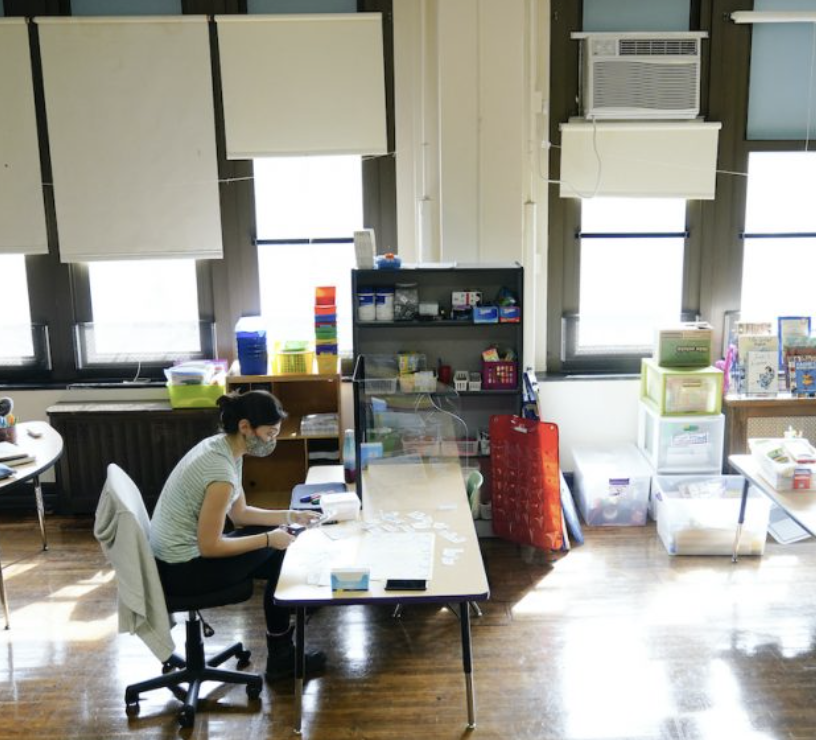 A teacher at Nebinger Elementary School in Philadelphia in 2021. The classroom has a big bank of window, a window unit air conditioner and shiny wooden floors.