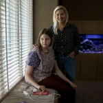 Devon Geiger, 22, and her mom, Deb. Devon sits in a window seat in a dim room. Deb stands with her hand on Devon's shoulder as Devon looks out the window.