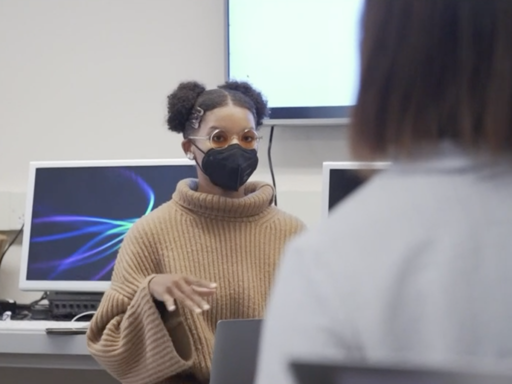 Screenshot from video shows a UC Berkeley student wearing a mask with hair in afro puffs speaking to someone blurry in the foreground.