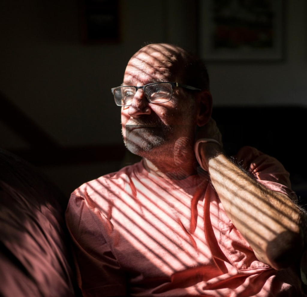 A man with light skin tone sits on a sofa looking out a window. The shades on the window cast shadows across his face.