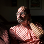 A man with light skin tone sits on a sofa looking out a window. The shades on the window cast shadows across his face.