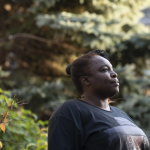 A woman with dark skin tone stands in front of trees and a brick building while looking off camera into the distance.