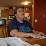 A middle-aged woman with long grey hair runs her hand over a paper while sitting in her kitchen.