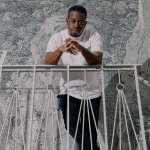 Zavier Sabio, who has dark toned skin and cropped curly hair, stands on a stairway with white iron railings in from of a wall covered in sketchy toile wallpaper