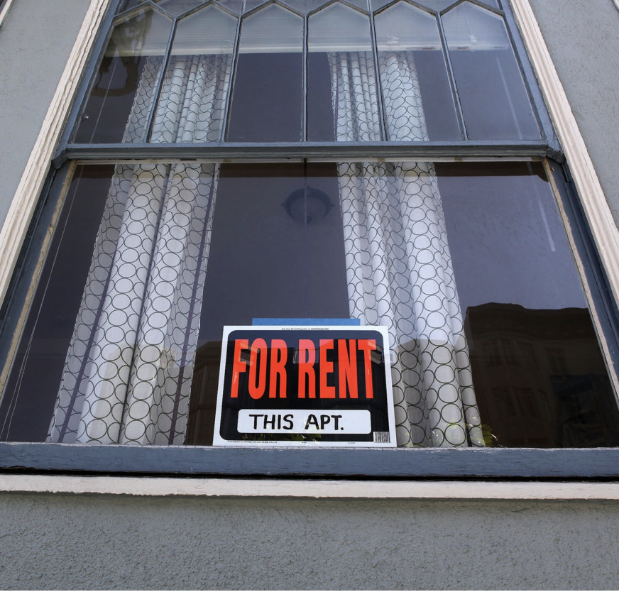 Leaded glass window in a grey stucco building with a For Rent sign posted.