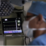 A person in blue scrubs and face mask stands in front of a vital signs monitor