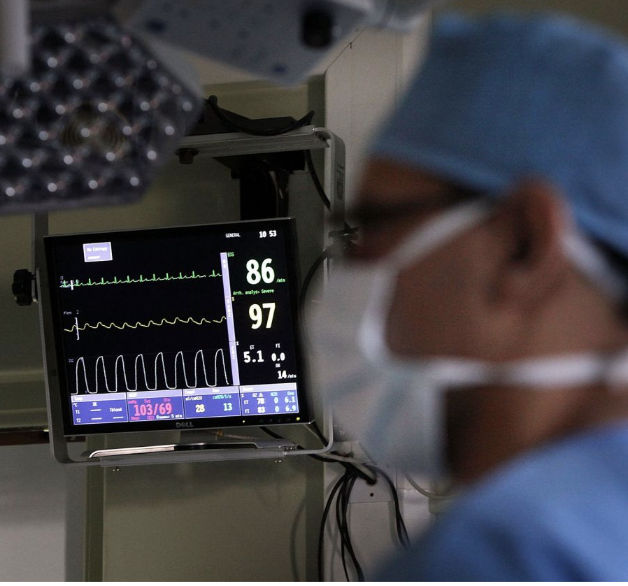 A person in blue scrubs and face mask stands in front of a vital signs monitor