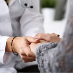 A person in a white lab coat with a stethoscope clasps hands with a person in a grey blouse.