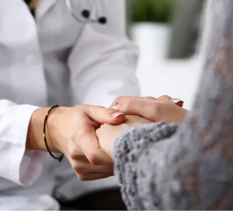 A person in a white lab coat with a stethoscope clasps hands with a person in a grey blouse.