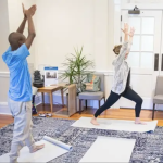 In a light-filled waiting room space, two people perform a standing yoga pose.