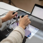 Light-toned hands manipulate a round device made of metal and plastic on a screen. Butterflies are shown on the screen.