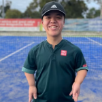 A young man with medium-toned skin and dwarfism stands on a blue tennis court.