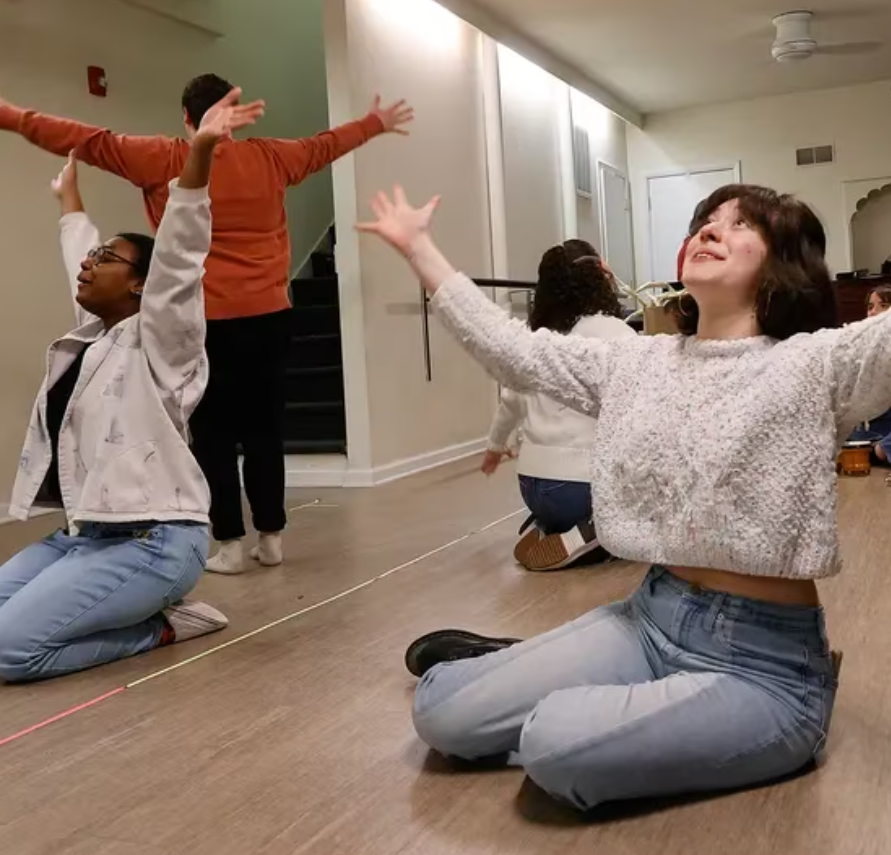 People in a dance studio sit on the floor or stand and spread their arms wide