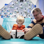 A child with light blond hair sits with their feet up and a box with buttons on their lap. An adult to the child's side points to a button on the box