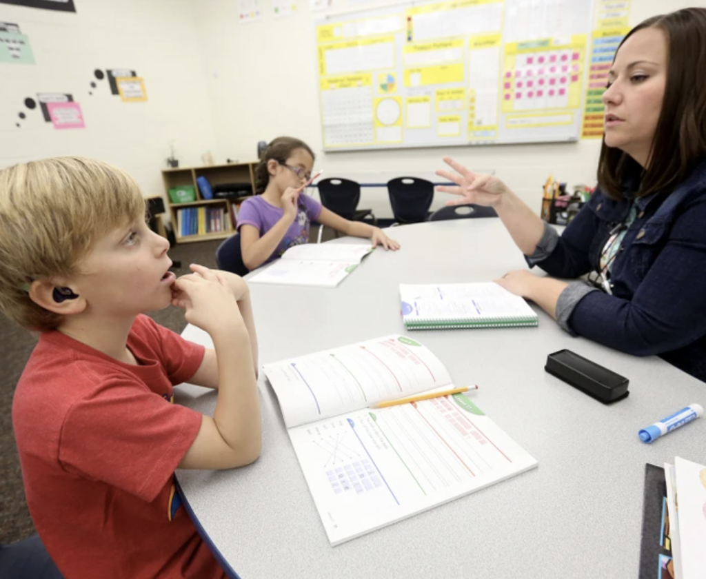 A teacher and young student sit across a classroom table from one another. The student has a hearing aie. The teacher holds three fingers out while speaking.