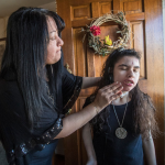 An adult with long dark hair wipes the face of a younger person with long dark hair at the door to a house.