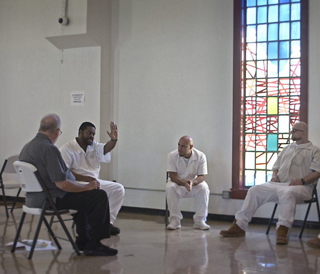 People in white jumpsuits sit on folding chairs with a person in street clothes in a room with large windows with some stained glass panels