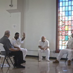 People in white jumpsuits sit on folding chairs with a person in street clothes in a room with large windows with some stained glass panels