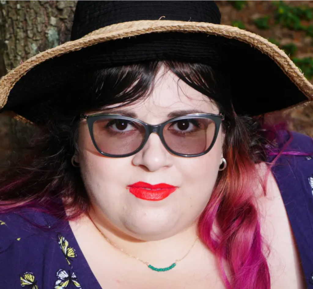A person with light-toned skin and dark and pink hair, a dainty turquoise necklace wears a hat, dark glasses and tangee lipstick.