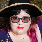 A person with light-toned skin and dark and pink hair, a dainty turquoise necklace wears a hat, dark glasses and tangee lipstick.
