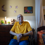 An older person sits in a wheelchair in a bedroom