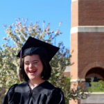 A young adult wearing black undergraduate academic regalia stands in front of a brick tower