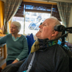 Bruce Goguen sits in a power wheelchair and Robin Goguen sits in a chair beside her husband. They are an older couple.