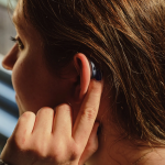 A young adult with long dark hair in profile. They wear a hearing aid and use a finger to adjust it.