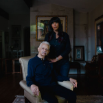 An adult daughter stands behind her aging father. Both have medium to dark toned skin.