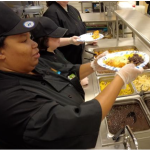 A Kitchen School student in black chef's coat plates food in a stainless steel kitchen.
