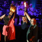 People gathered at night. Two hold up a ceremonial torch. One holds up a championship belt.