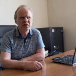 A person in a polo shirt sitting by a laptop with a safe in the background.