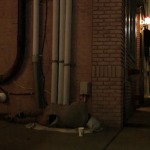 A dark street. A person sleeps on the ground next to a brick building.