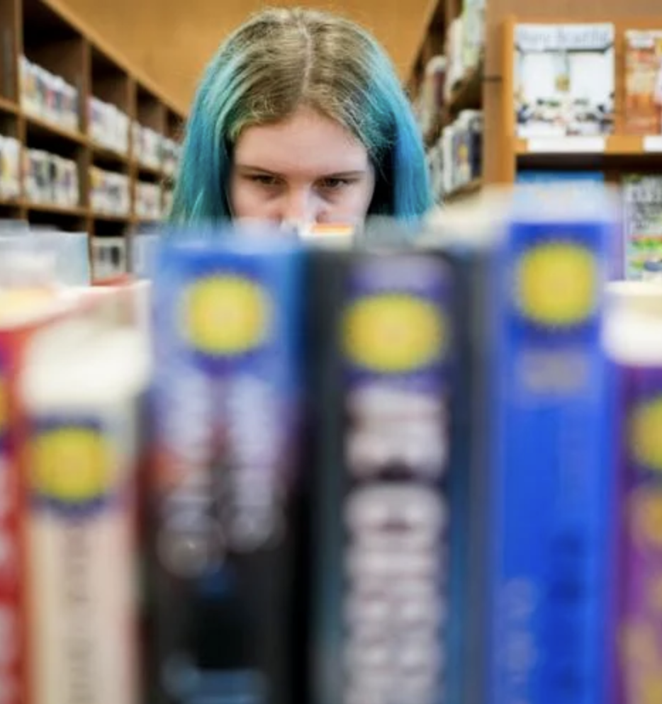 A person with blue hair seen beyond a shelf of books.