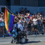 A person in a power wheelchair carries a rainbow flag by a street side crowd