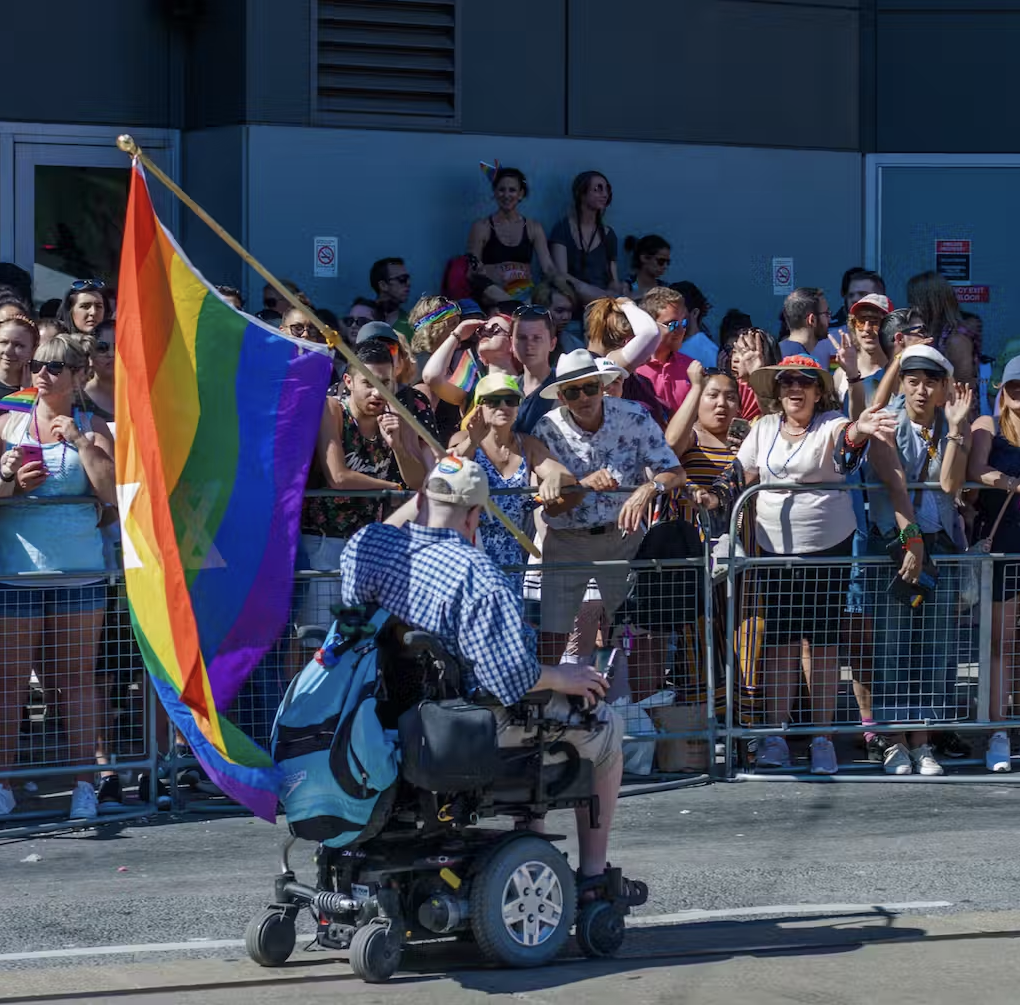 A person in a power wheelchair carries a rainbow flag by a street side crowd