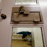 A metal door with a lock, small windows and system for delivering meals looks into a room with a cot , tile floor and toilet.