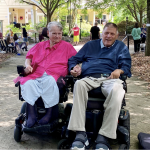 Eleanor Smith sits in a power chair, holding hands with Mark Johnson, also seated in a power chair