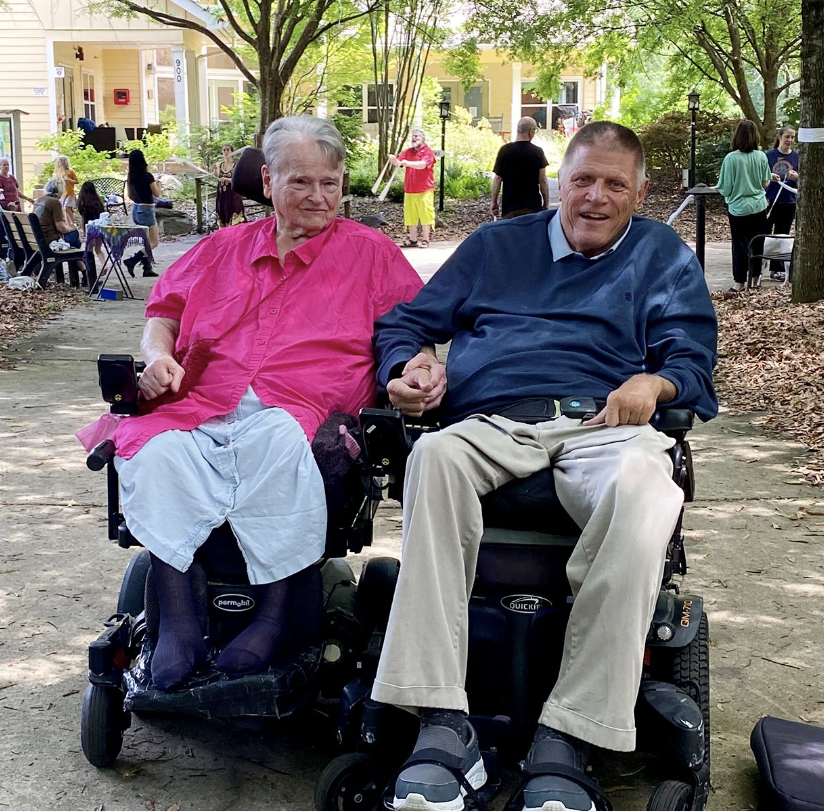 Eleanor Smith sits in a power chair, holding hands with Mark Johnson, also seated in a power chair