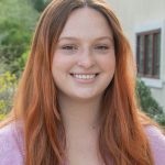 Headshot of Hannah Mennella. Hannah is an adult female with long auburn hair and is wearing a pink sweater.