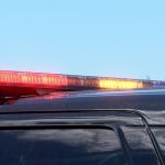 Close-up of a police vehicle’s roof with red and amber emergency lights illuminated against a clear blue sky. A streetlight and security camera are visible in the background.