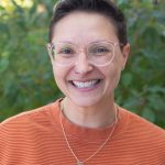 Headshot of Jess Monahan. Jess has short brown hair and glasses and is wearing an orange ribbed sweater.