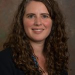 Headshot of Sarah Curtiss. Sarah has long brown curly hair and brown eyes. She is wearing a black blazer.