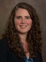Headshot of Sarah Curtiss. Sarah has long brown curly hair and brown eyes. She is wearing a black blazer.